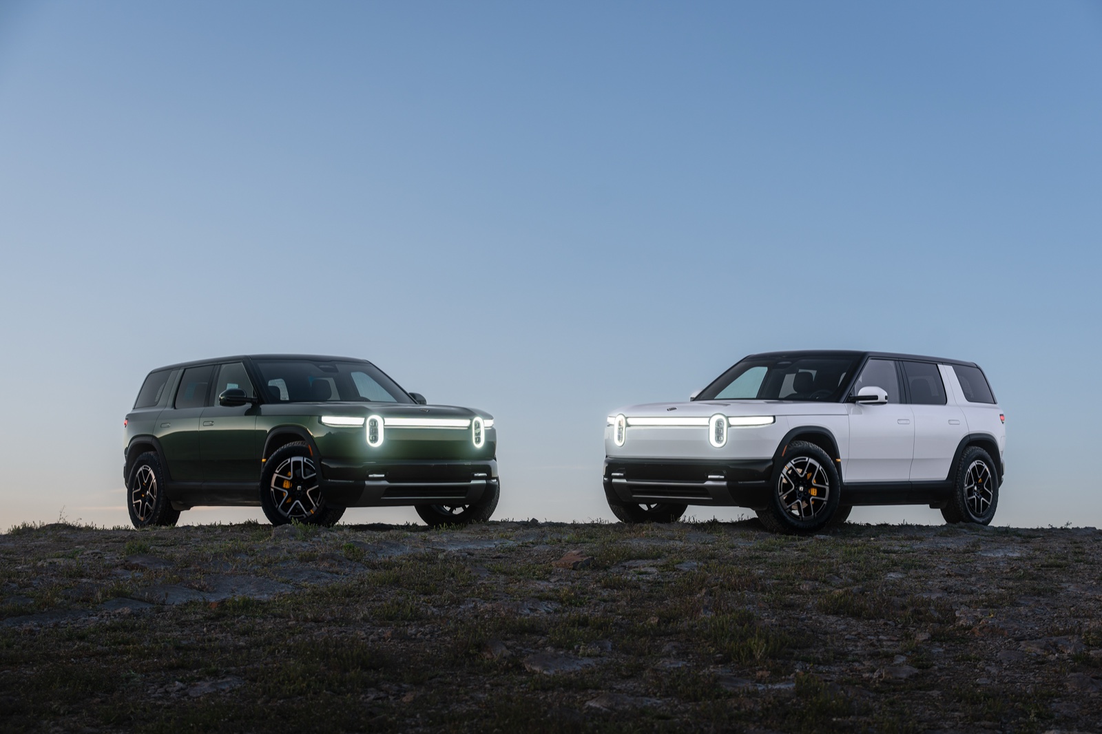 Dos SUV eléctricos Rivian R2 en verde y blanco estacionados uno al lado del otro en una cima rocosa al anochecer